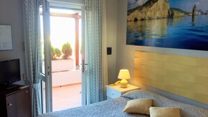 A hotel room with a bed, a nightstand with a lamp, a painting of a coastal scene, and a view of the outdoors through a door. - Hotel Ortensia (Ponza)