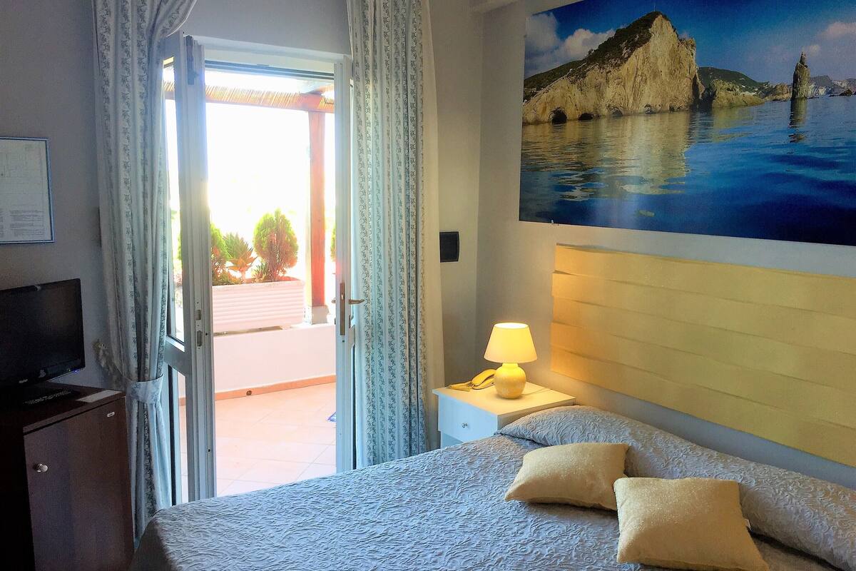 Une chambre d’hôtel avec un lit, une table de chevet avec une lampe, un tableau représentant un paysage côtier et une vue sur l’extérieur par une porte.