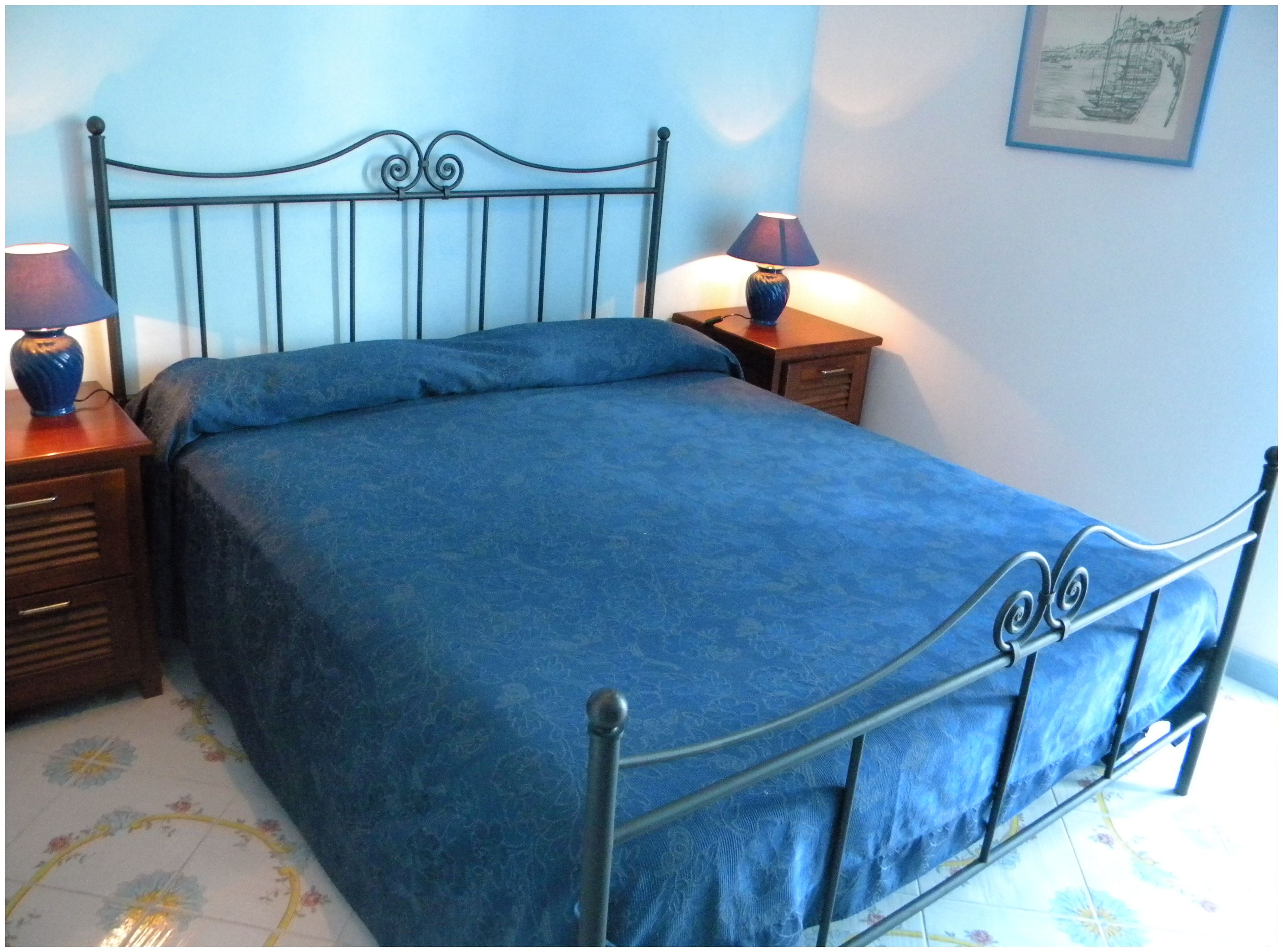 Ein Bett mit Metallgestell und blauer Bettdecke, flankiert von zwei Nachttischen mit Lampen, in einem Raum mit Fliesenboden.