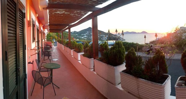 A balcony with outdoor seating, potted plants, and a view of the sea and hills at sunset. - Hotel Ortensia (Ponza)
