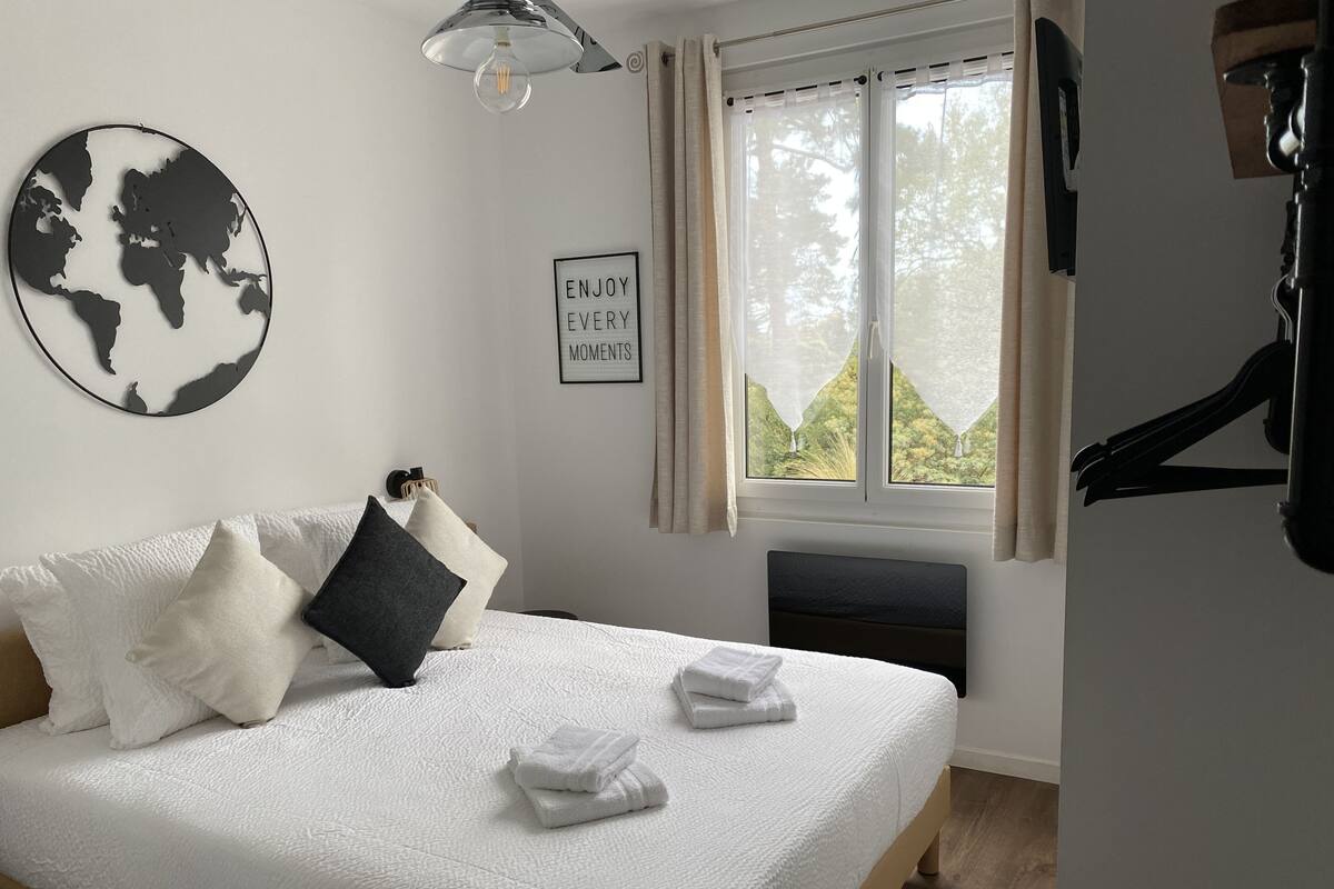 A neatly made bed with white bedding and two towels, a world map artwork on the wall, a framed inspirational quote, and a window with curtains.