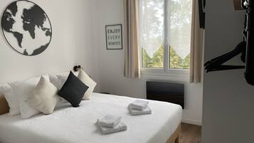 Een netjes opgemaakt bed met witte beddengoed en twee handdoeken, een wereldkaart als kunstwerk aan de muur, een ingelijst inspirerend citaat, en een raam met gordijnen.