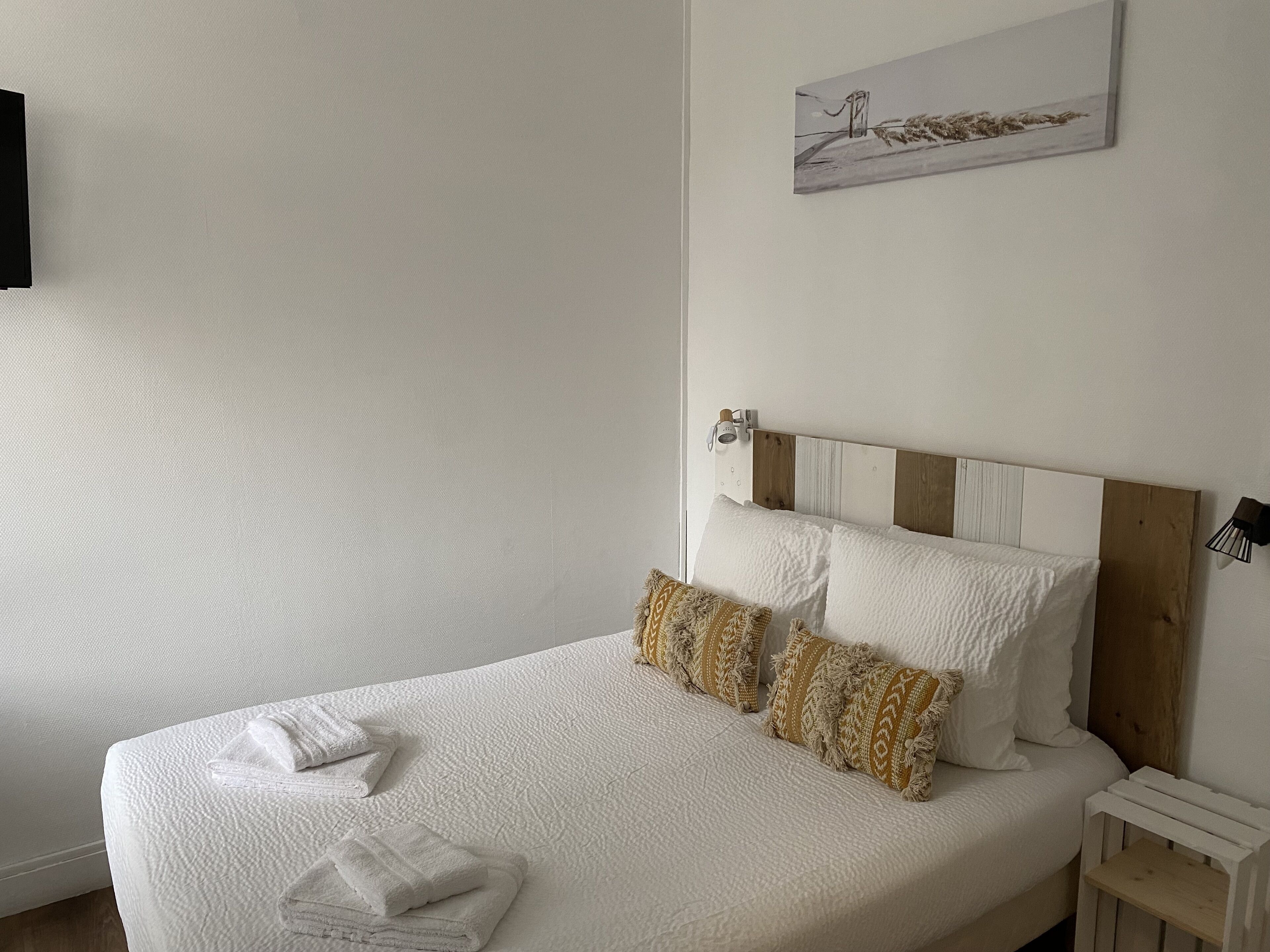 Una cama bien tendida con ropa blanca y almohadas estampadas, una mesita de noche pequeña y una lámpara fijada a la pared.