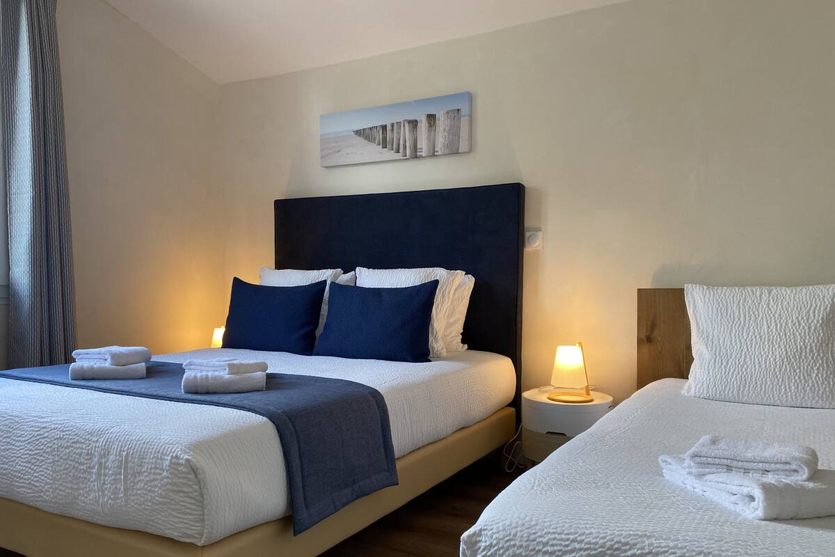 A hotel room with two beds, a headboard, a bedside table with a lamp, and a painting on the wall.