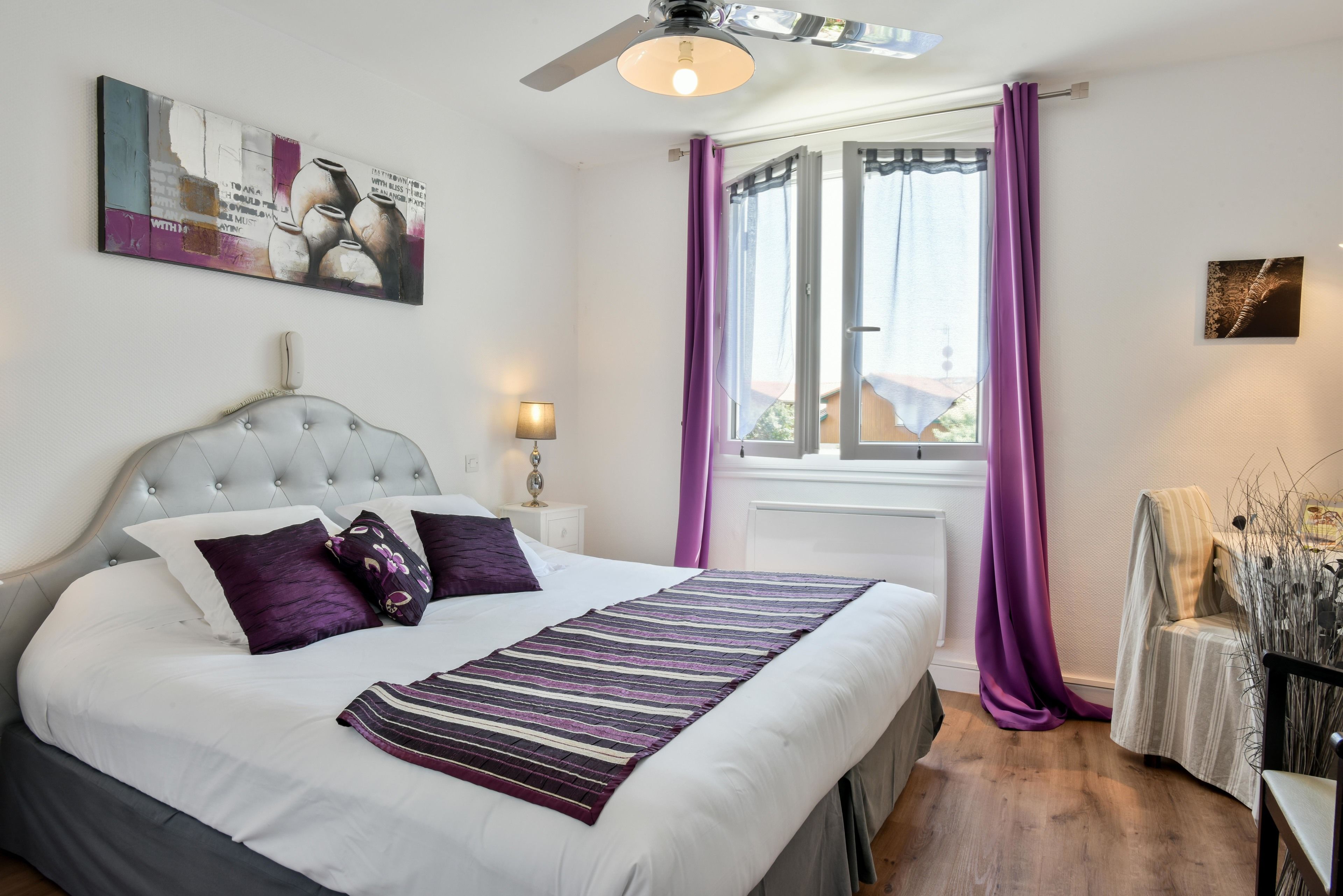 Een slaapkamer met een groot bed, een plafondventilator, een raam met paarse gordijnen en een kunstwerk aan de muur.