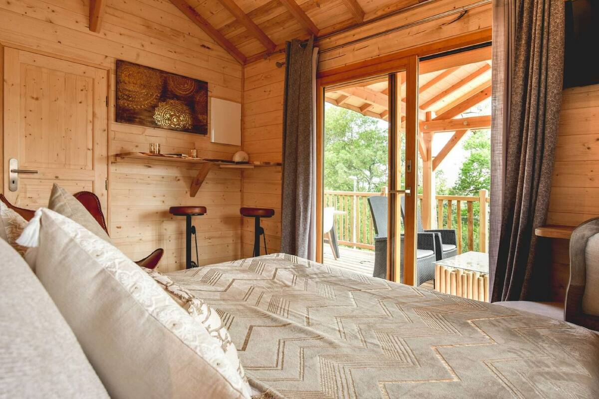 A wooden cabin room with a bed, a small table, two stools, and a balcony with outdoor furniture.
