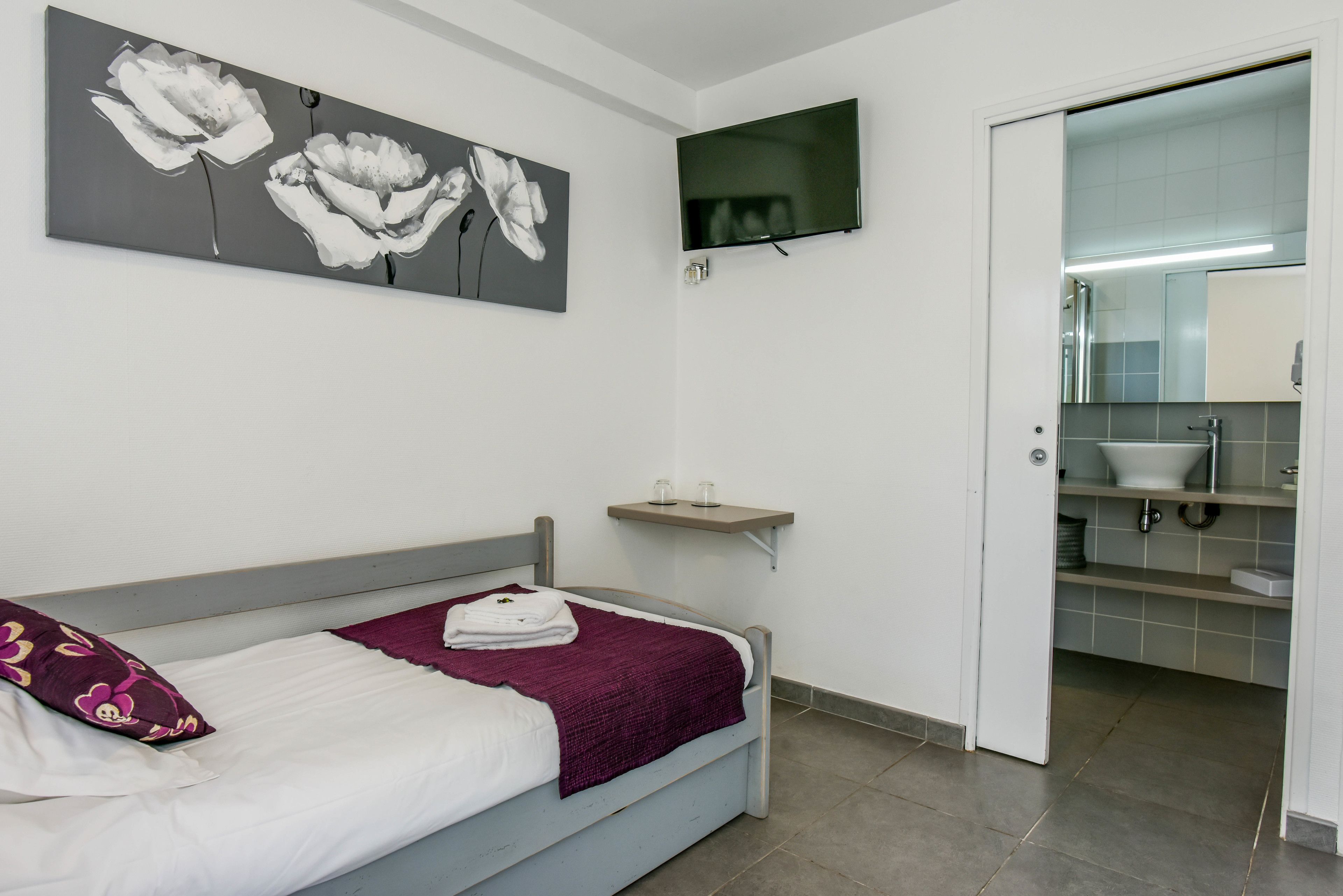 A bedroom with a bed, a TV mounted on the wall, a bathroom visible through an open door, and a shelf with two glasses.
