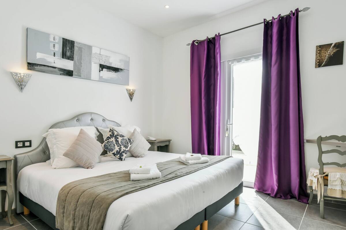 A neatly made bed with a grey headboard, white bedding, and a grey throw blanket. There are two bedside tables with lamps, a wall-mounted artwork, and a window with purple curtains.