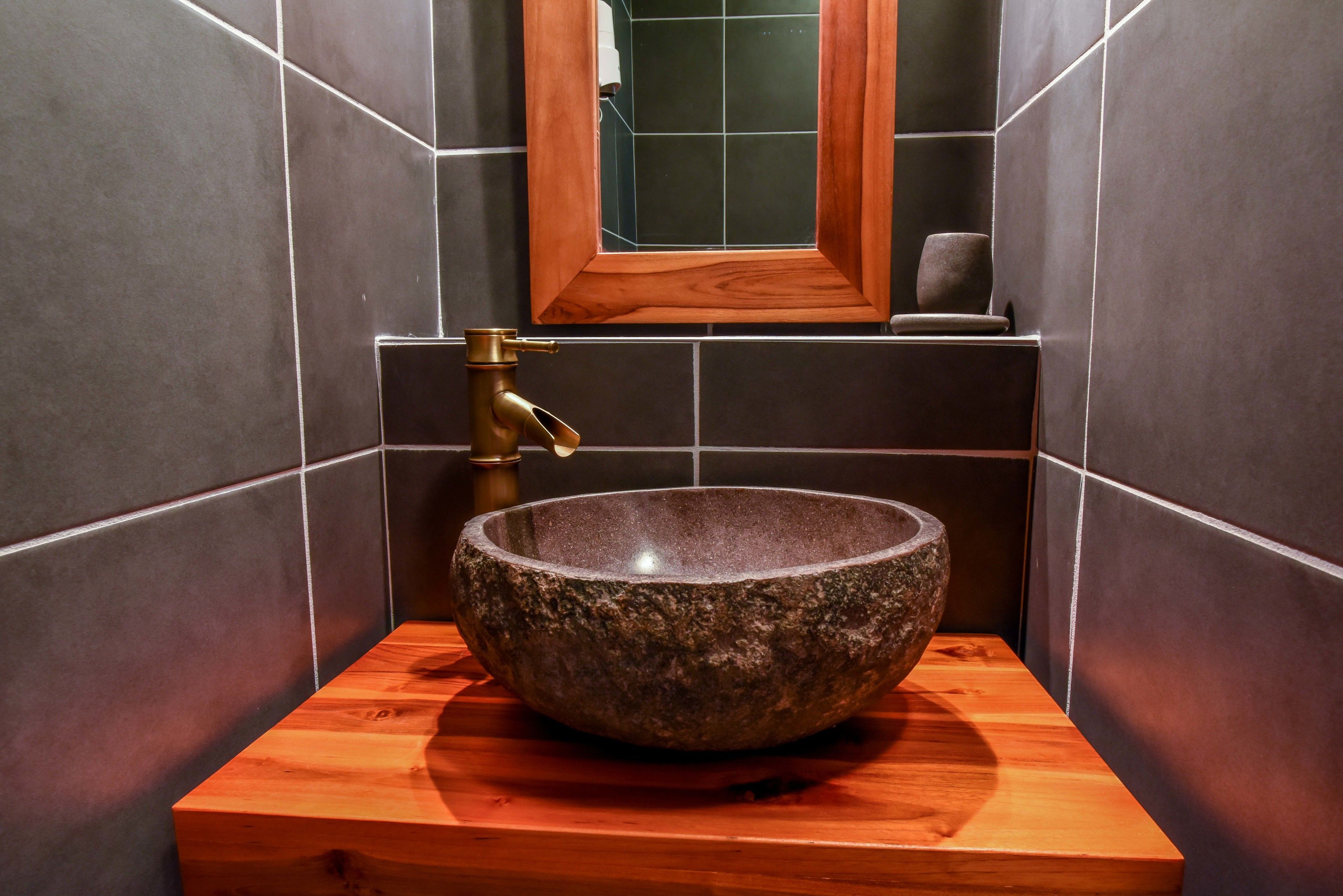 A stone sink with a wooden base in a bathroom with dark tiles and a wooden-framed mirror.