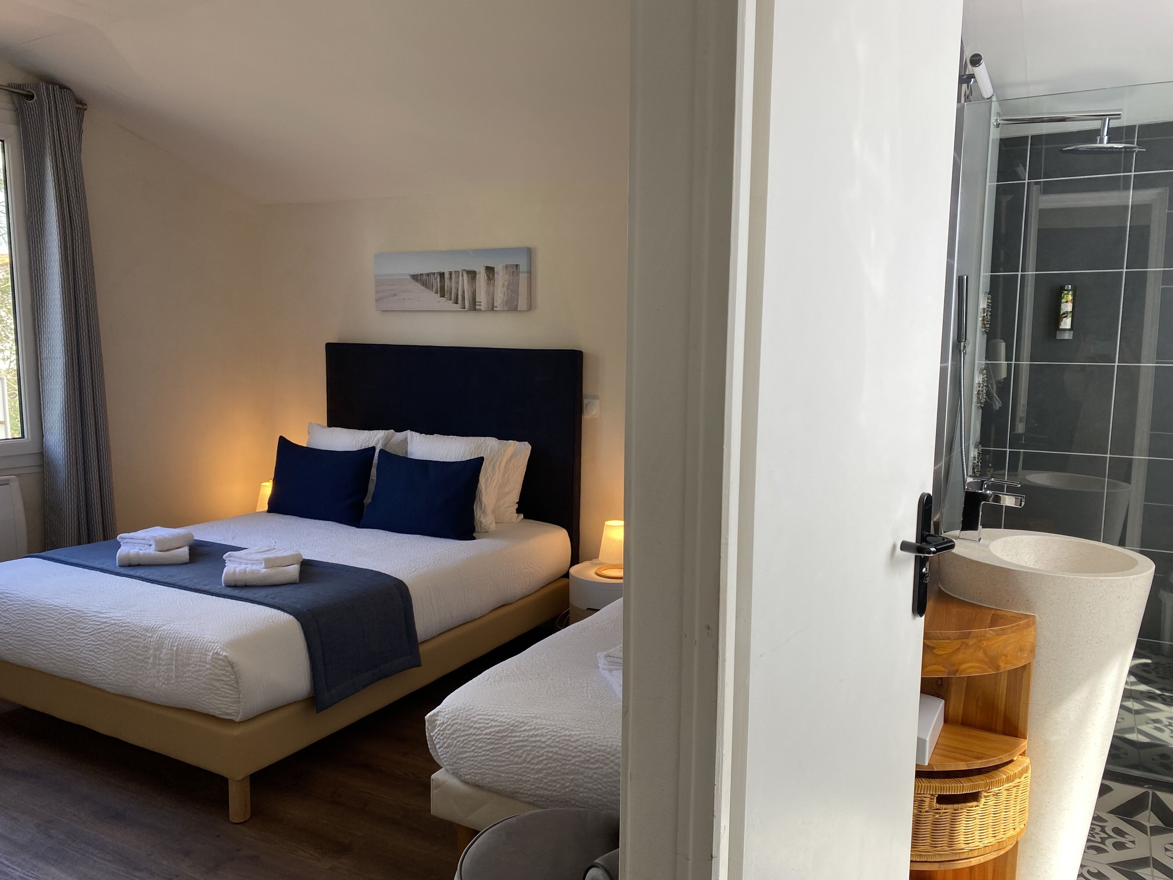 Une chambre d’hôtel moderne avec un grand lit, une table de chevet, une petite chaise, une salle de bain avec une douche en verre et une table d’appoint en bois.