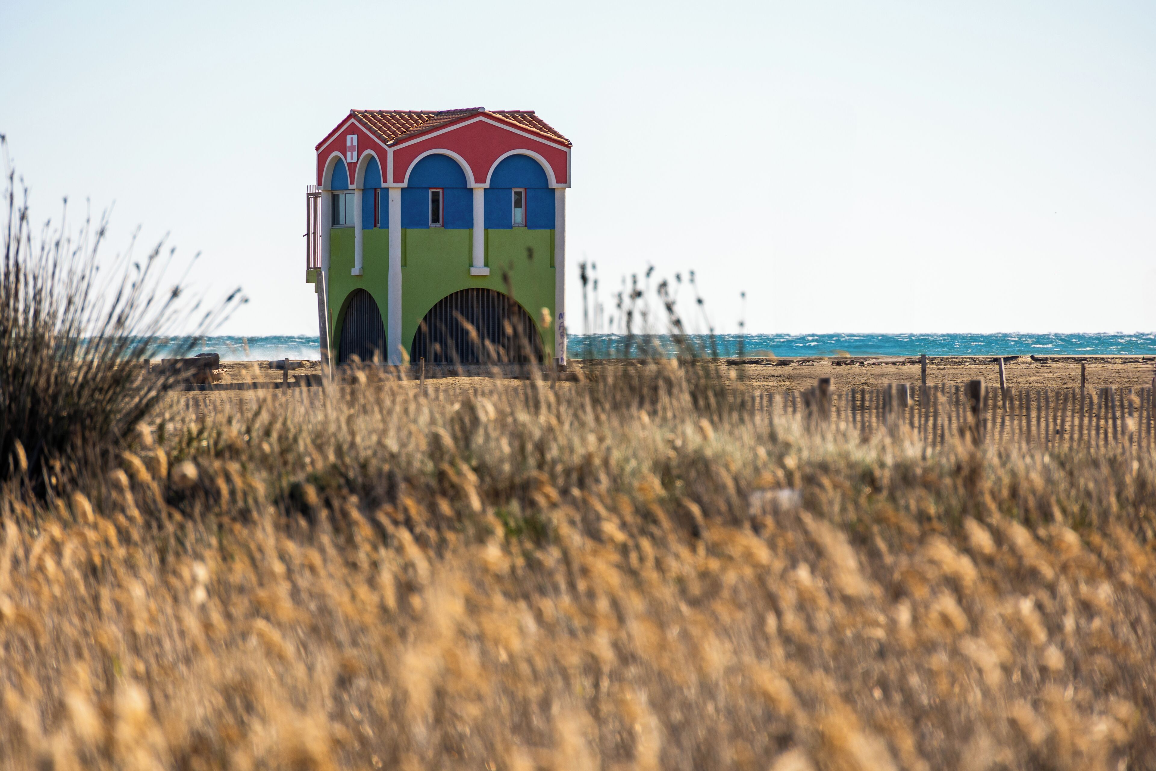 Un edificio colorido con techo rojo y paredes azules y verdes, situado en una playa con hierba seca al frente.