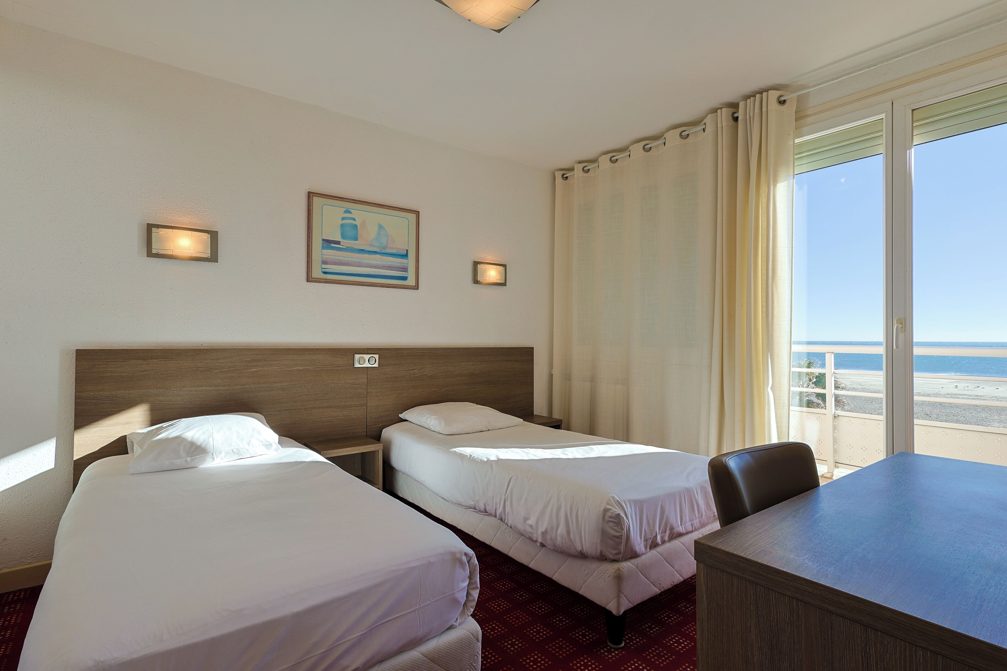 A hotel room with two single beds, a desk, and a view of the beach.