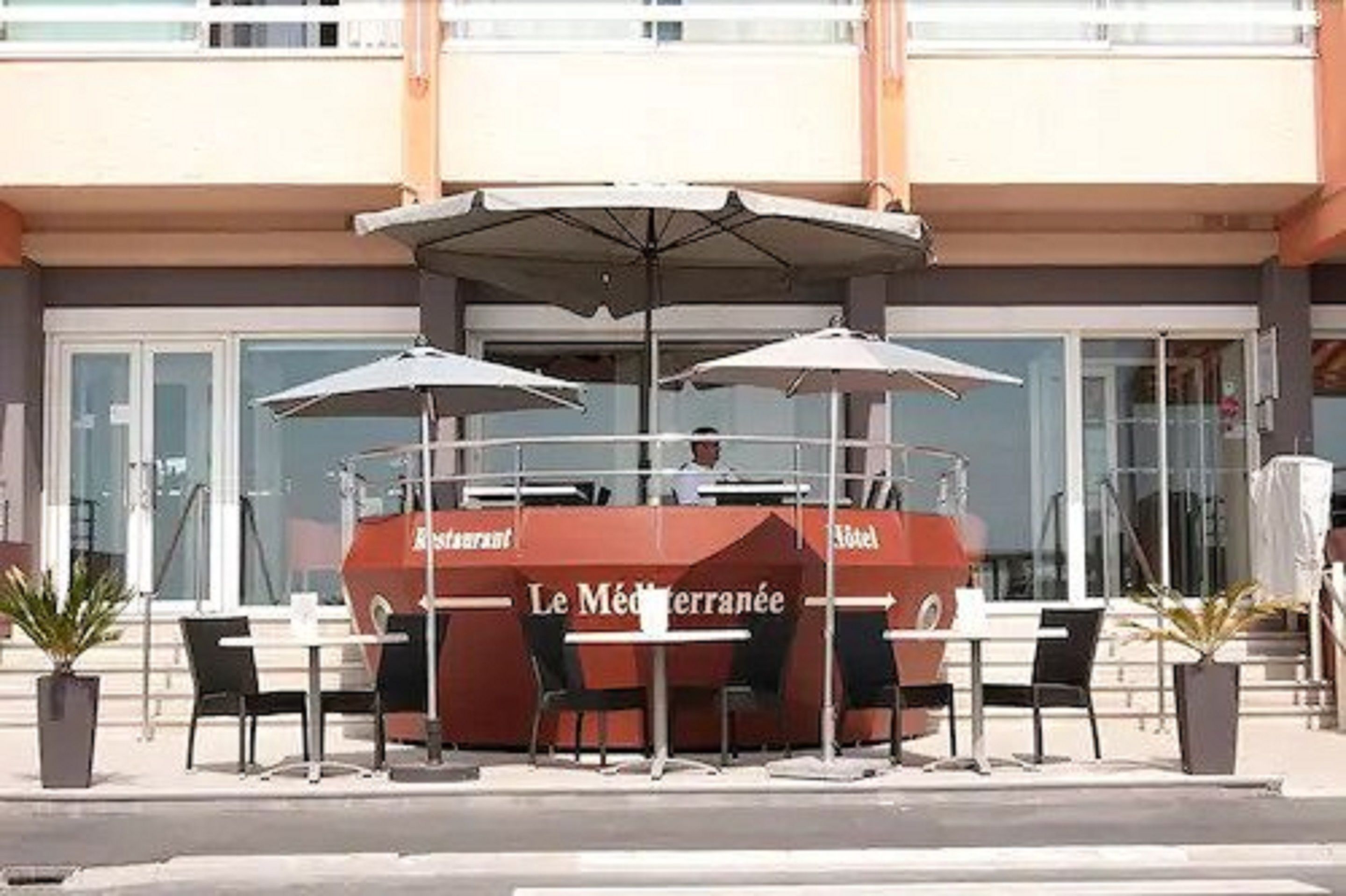 Un espace extérieur aménagé avec des tables et des parasols, une enseigne indiquant « Le Méditerranée », et l’entrée d’un bâtiment en arrière-plan.