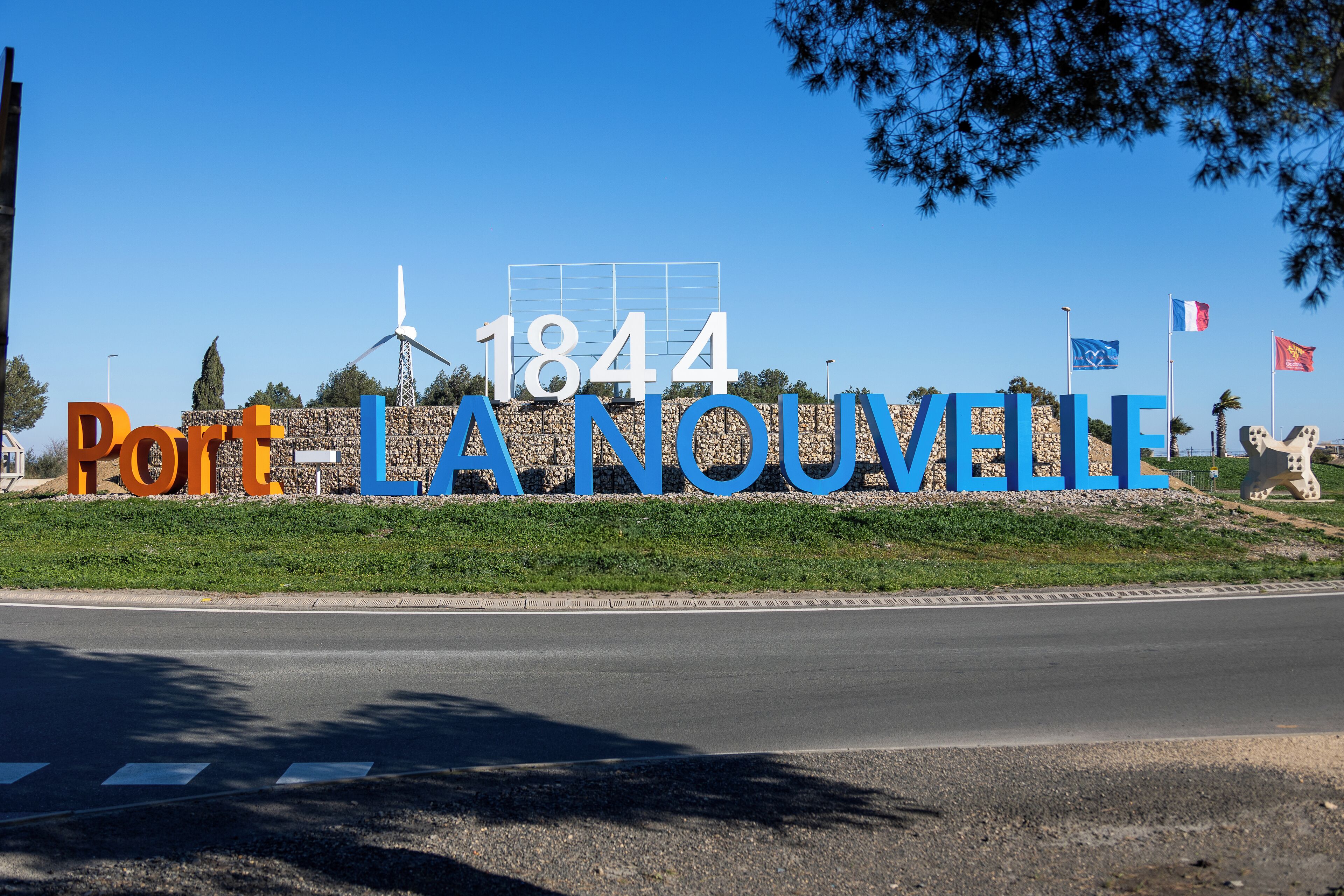 Un letrero con el año 1844 y las palabras "Port-LANOUVELLE" en letras grandes, con banderas y una turbina eólica al fondo.