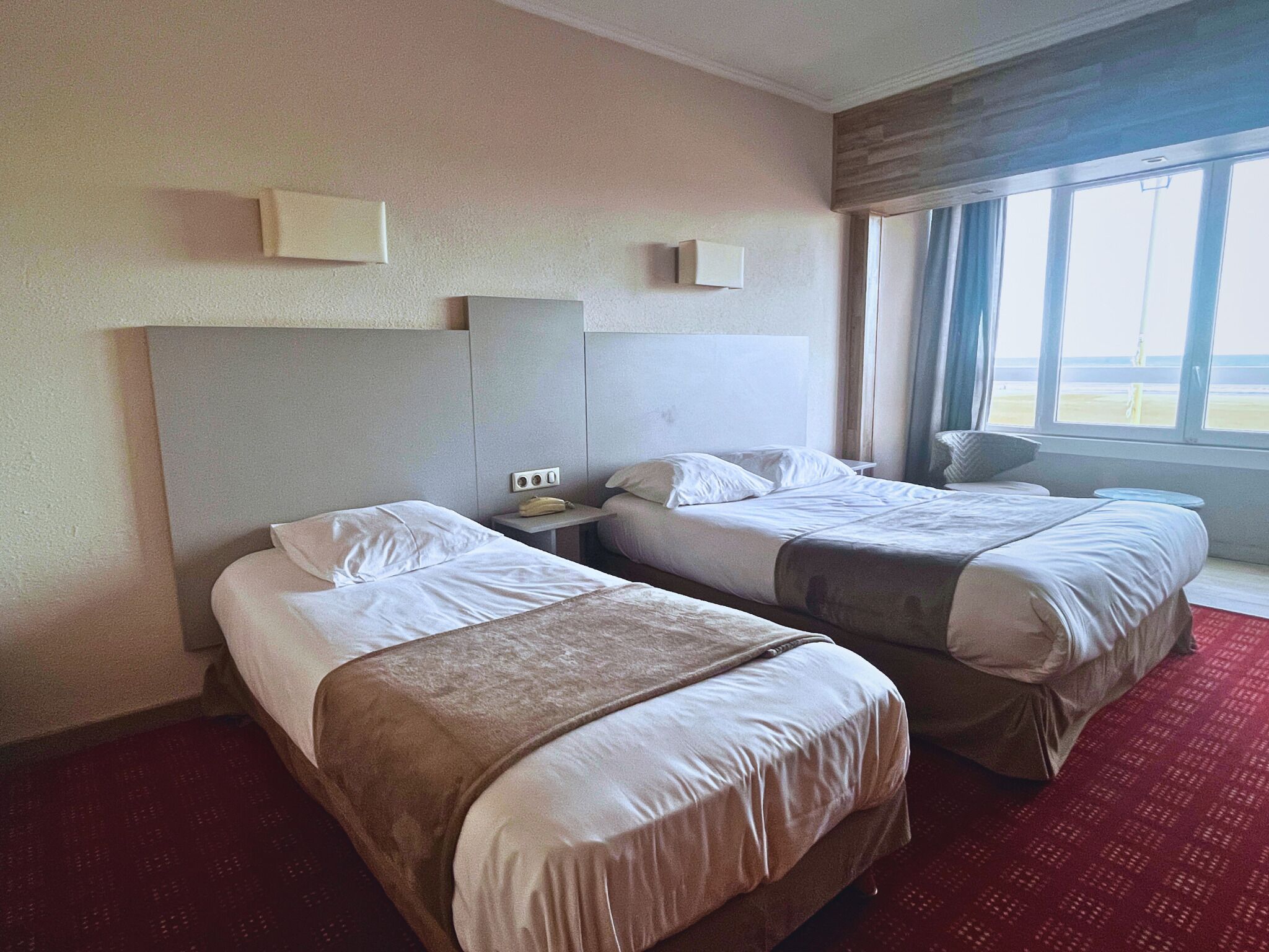A hotel room with two beds, a small table, and a window with a view.