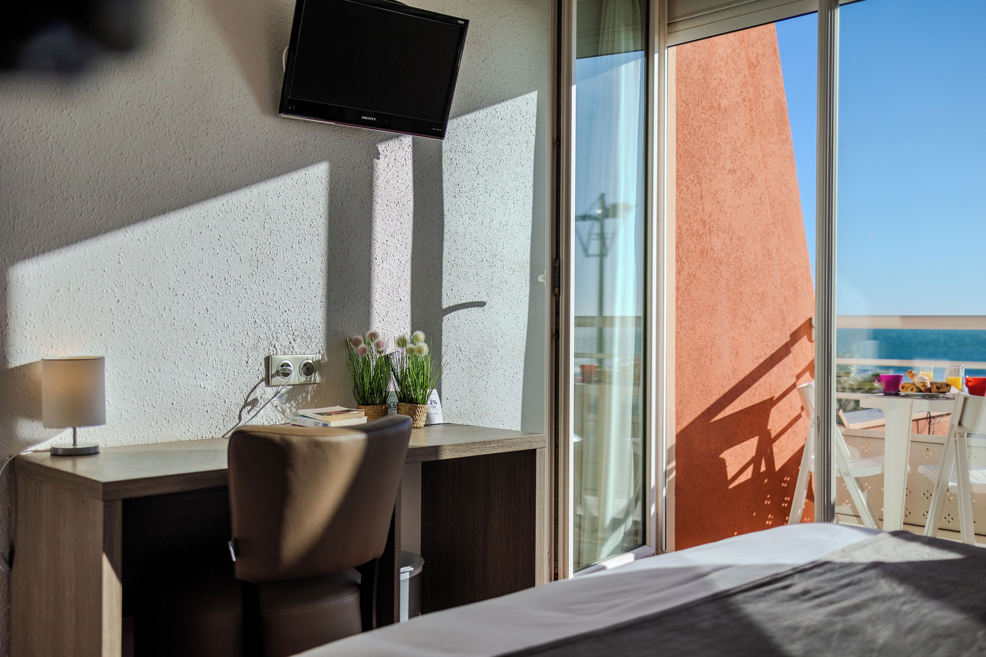 A hotel room with a bed, a desk with a chair, a television mounted on the wall, a small table with a lamp, and a window with a view of the sea.