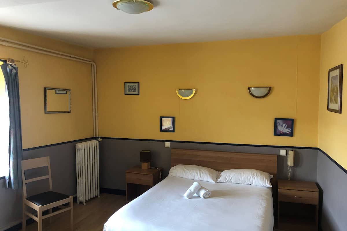 Habitació d'hotel amb un llit, una cadira, una taula de nit, una làmpada i un radiador.