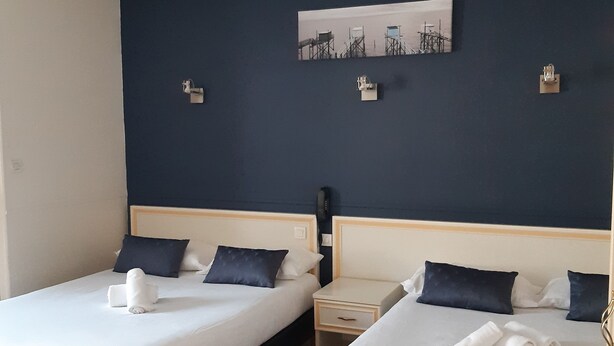 Quarto de hotel com duas camas, cada uma com roupa de cama branca e almofadas escuras, uma pequena mesa de cabeceira com abajur e um quadro na parede.