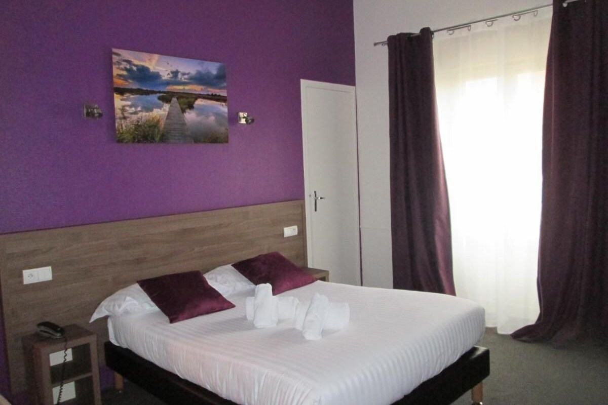 Un lit bien fait, avec du linge de lit blanc et des coussins violets, une tête de lit en bois, une table de chevet avec un téléphone, et un tableau représentant un coucher de soleil sur un pont, accroché au mur.