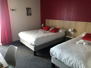 A hotel room with two beds, red pillows, a small table, and a chair.