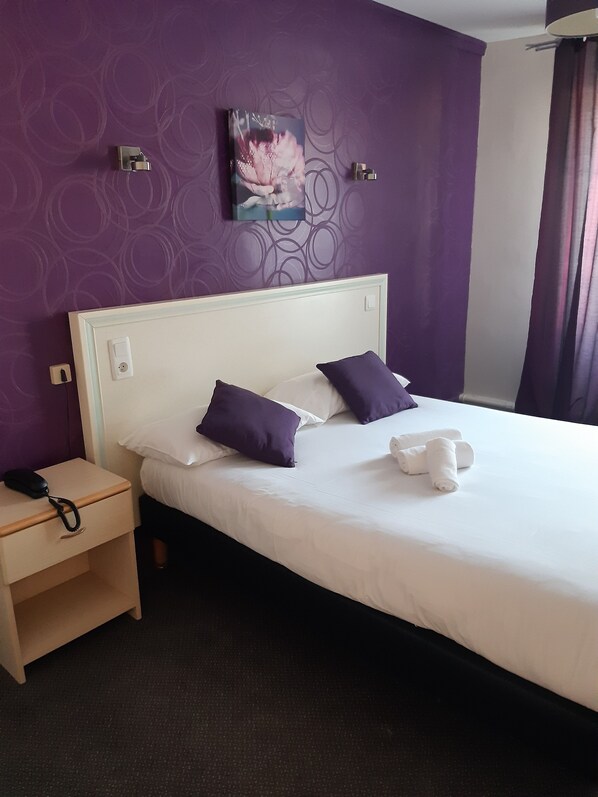 A neatly made bed with white and purple bedding, a nightstand with a phone, and a painting on the wall. - Hostellerie du Cheval Blanc (Sainte-Maure-de-Touraine)
