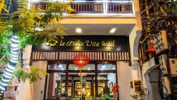 A multi-story building with balconies, a sign reading "La Dolce Vita Hotel," and outdoor seating with potted plants.