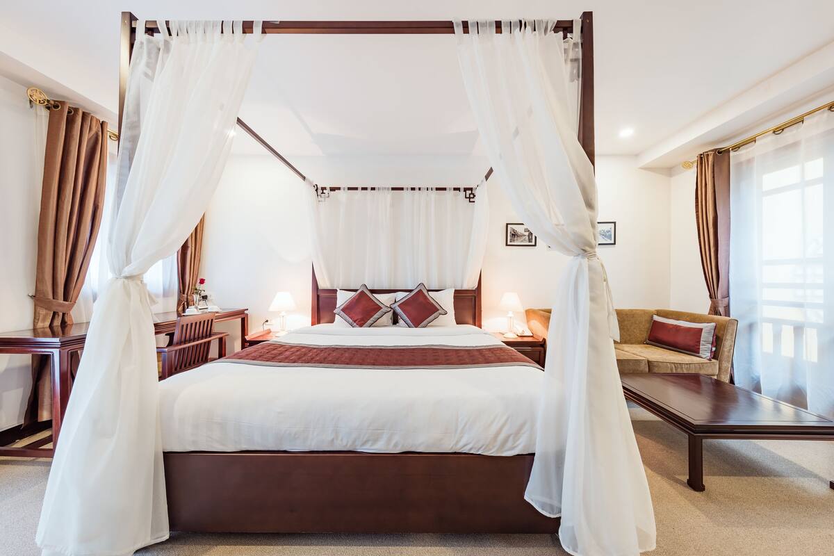 A four-poster canopy bed with a wooden frame, a large bed with a red and white bedspread, two bedside tables with lamps, a sofa with a red cushion, and a picture on the wall.