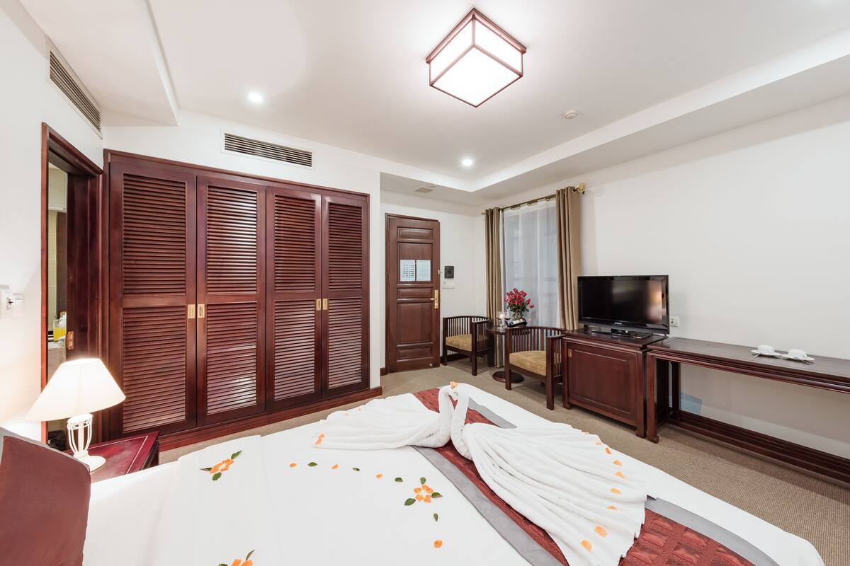 A hotel room with a large bed, a desk with a television, a wardrobe, and a small table with a lamp.