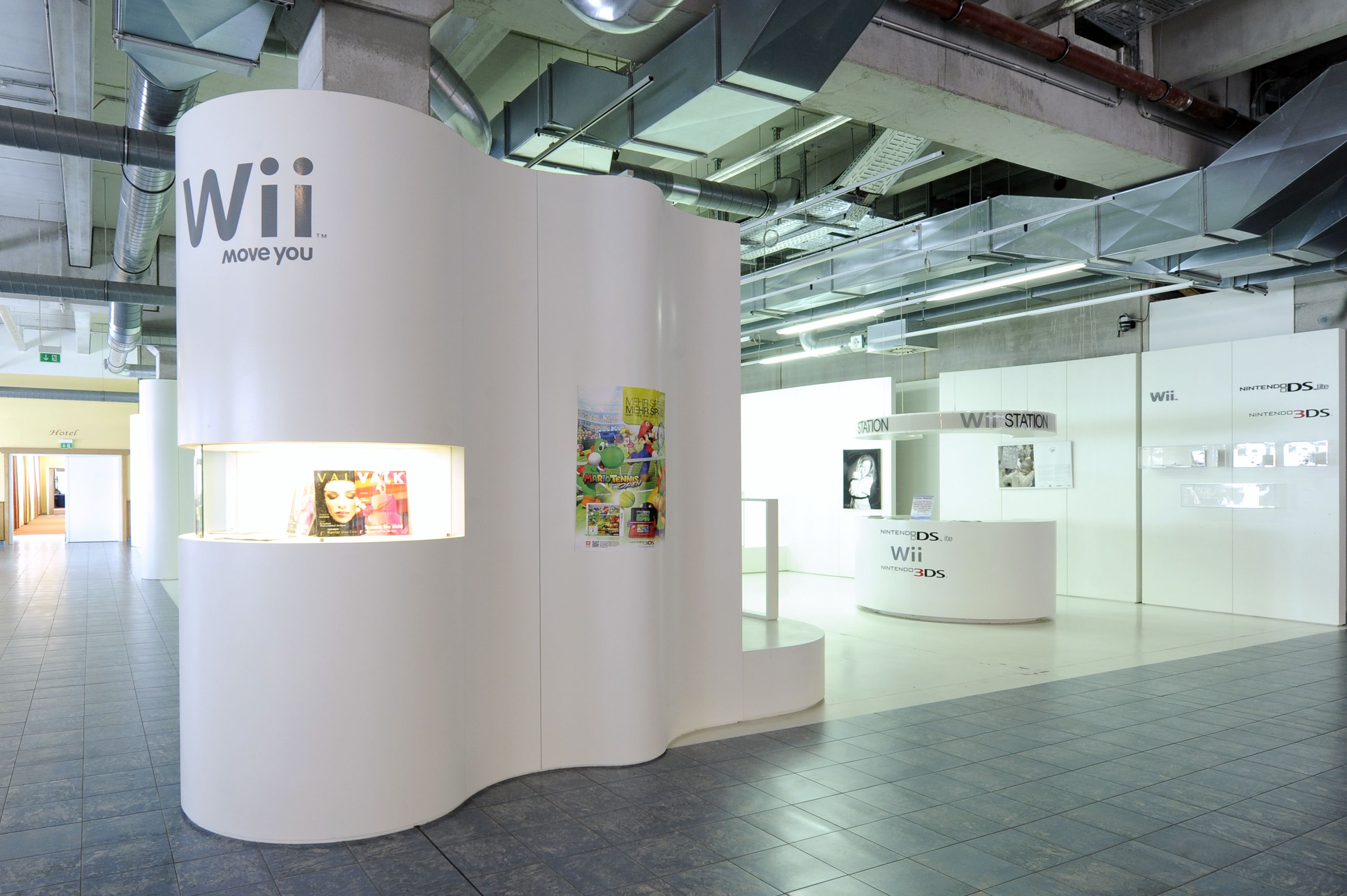 Un quiosco moderno y blanco con la marca 'Wii Move You', que incluye un mostrador de revistas y carteles promocionales.