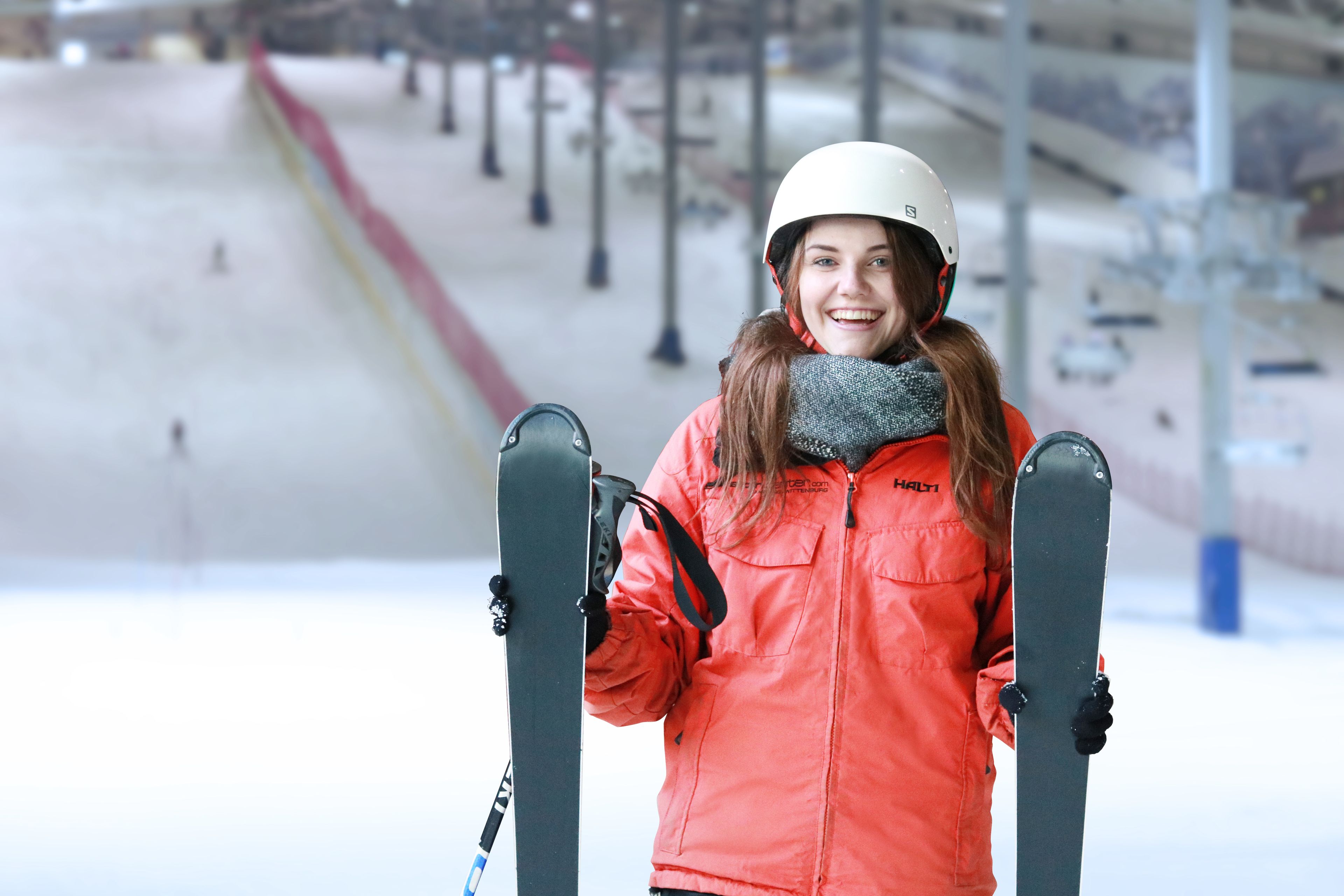 A person in a red jacket holding skis, standing on a snowy surface with a ski lift in the background.