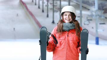 Una persona con chaqueta roja sosteniendo esquíes, de pie sobre nieve, con un telesilla al fondo.