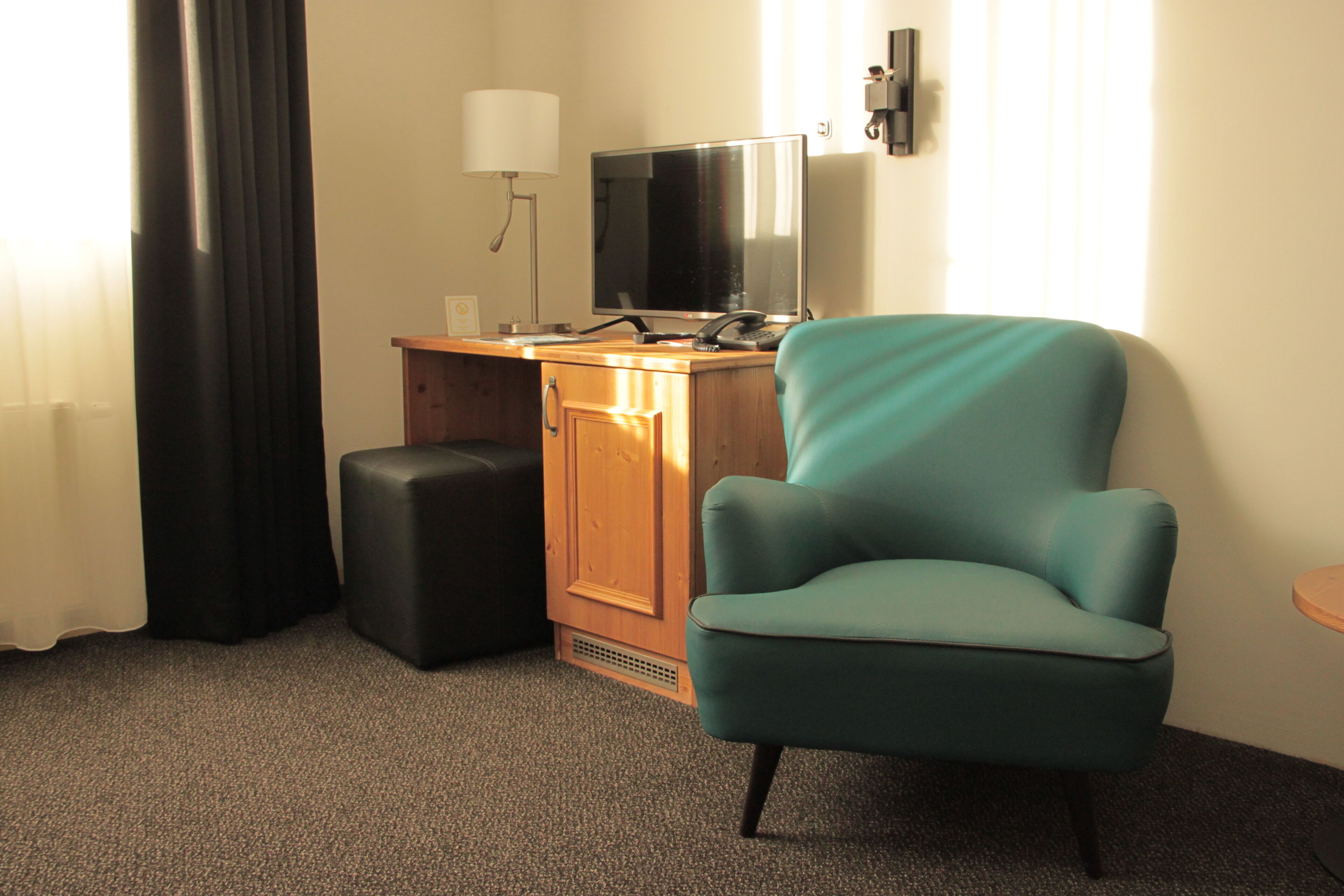Een hotelkamer met een blauwgroene fauteuil, een houten bureau met een televisie, een lamp en een zwart poef.