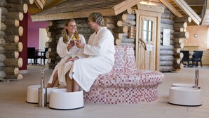 A couple in white robes sitting on a mosaic-patterned bench in a log cabin-style room with wooden beams and a stone wall.