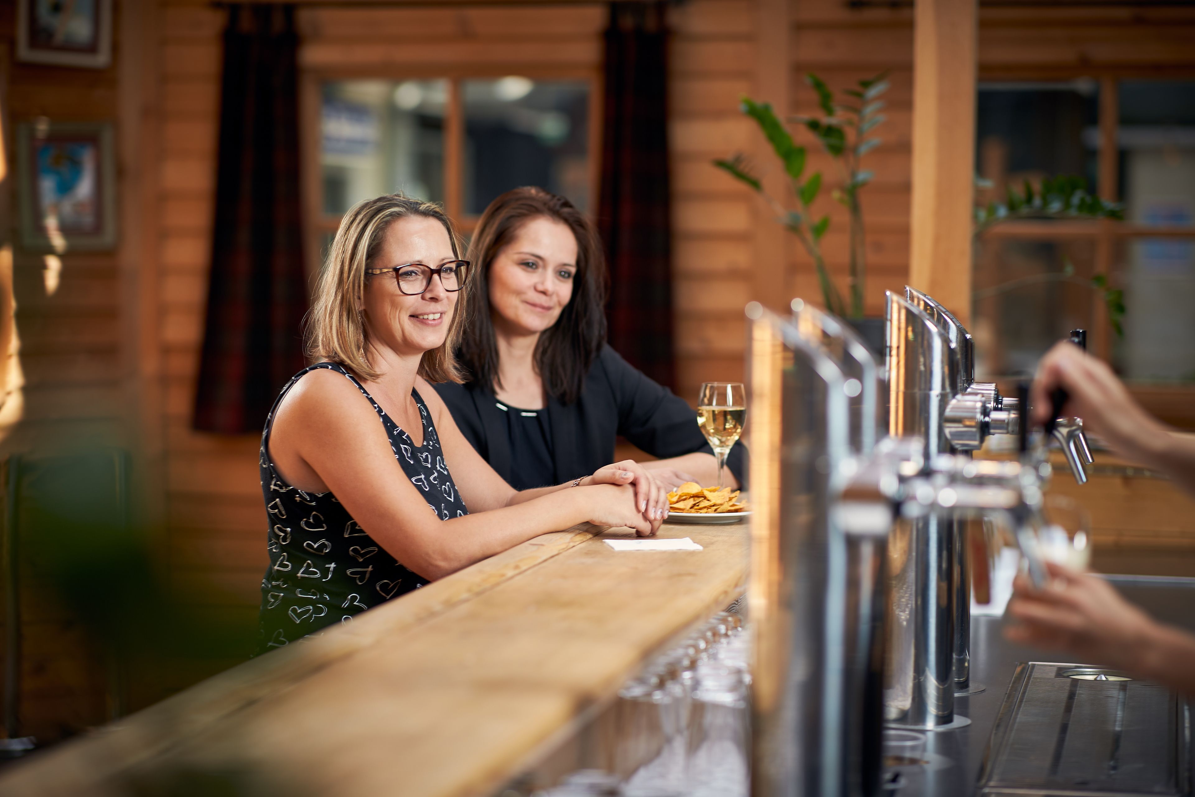Twee personen aan een bar met een wijnglas en een bord met hapjes.
