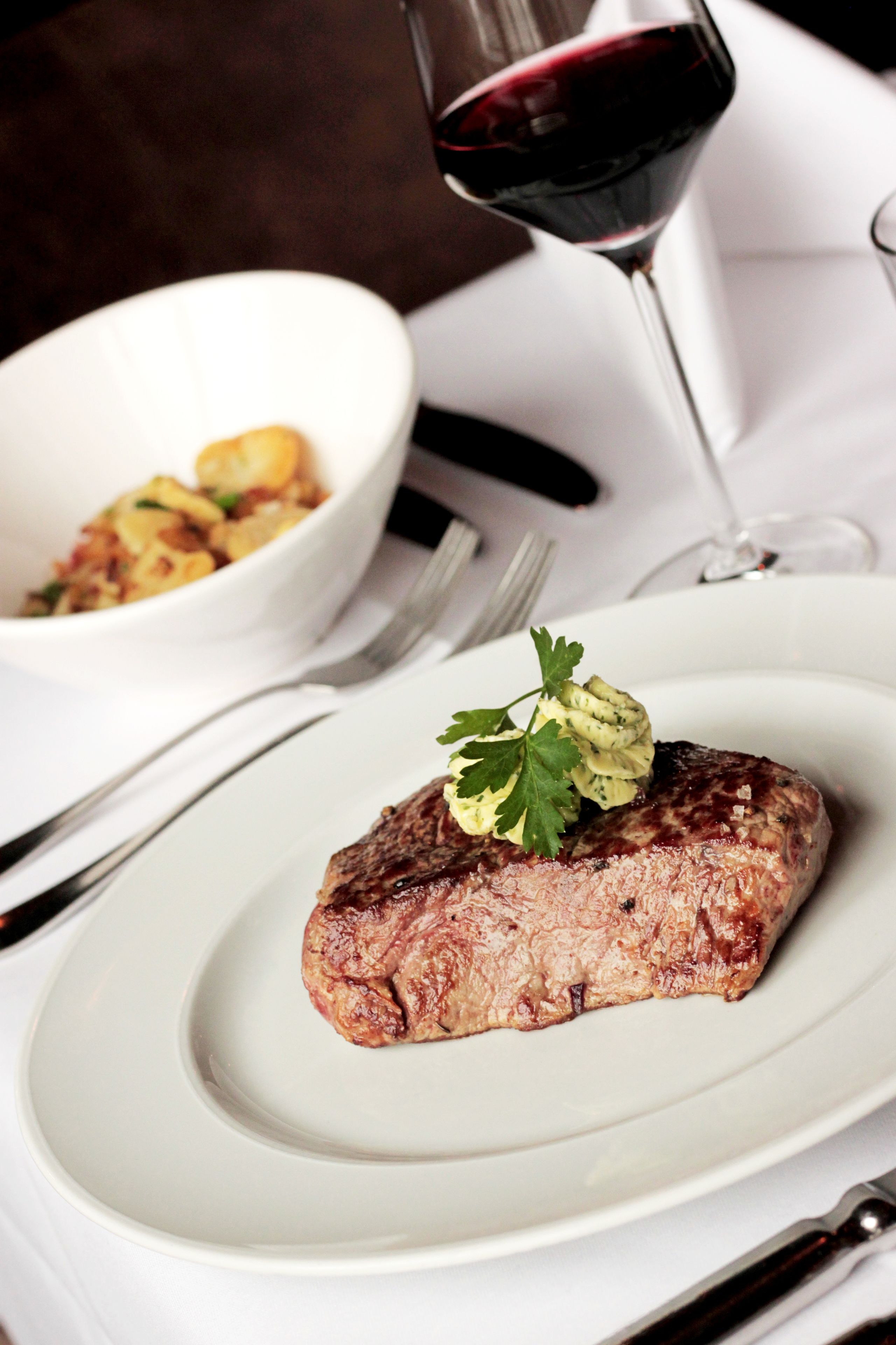 A steak with a garnish on a plate, a glass of red wine, and a bowl of food in the background.