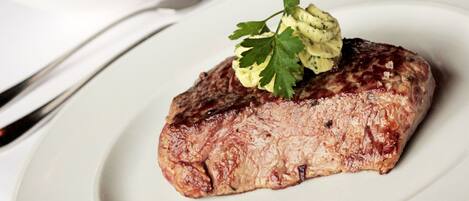 A steak with a garnish on a plate, a glass of red wine, and a bowl of food in the background.