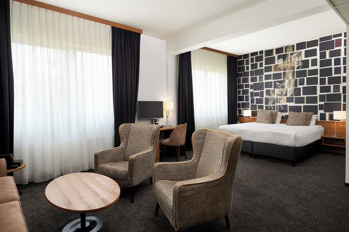 Ein Hotelzimmer mit einem Bett, zwei Sesseln, einem runden Tisch, einem Schreibtisch und einer Wand mit geometrischem Muster.