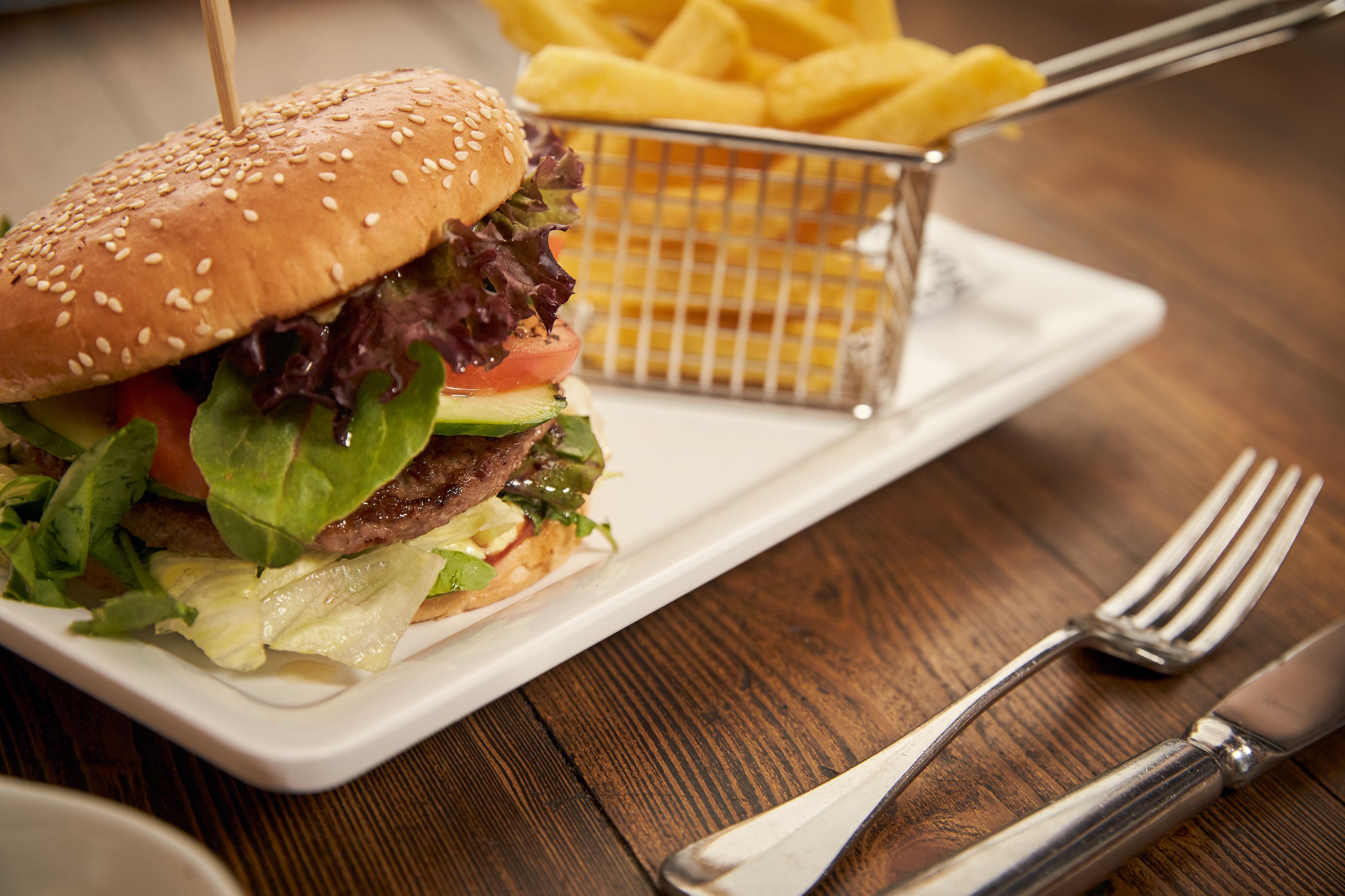 A burger with lettuce, tomato, and a sesame seed bun on a white plate, accompanied by a basket of fries and a fork on a wooden table.