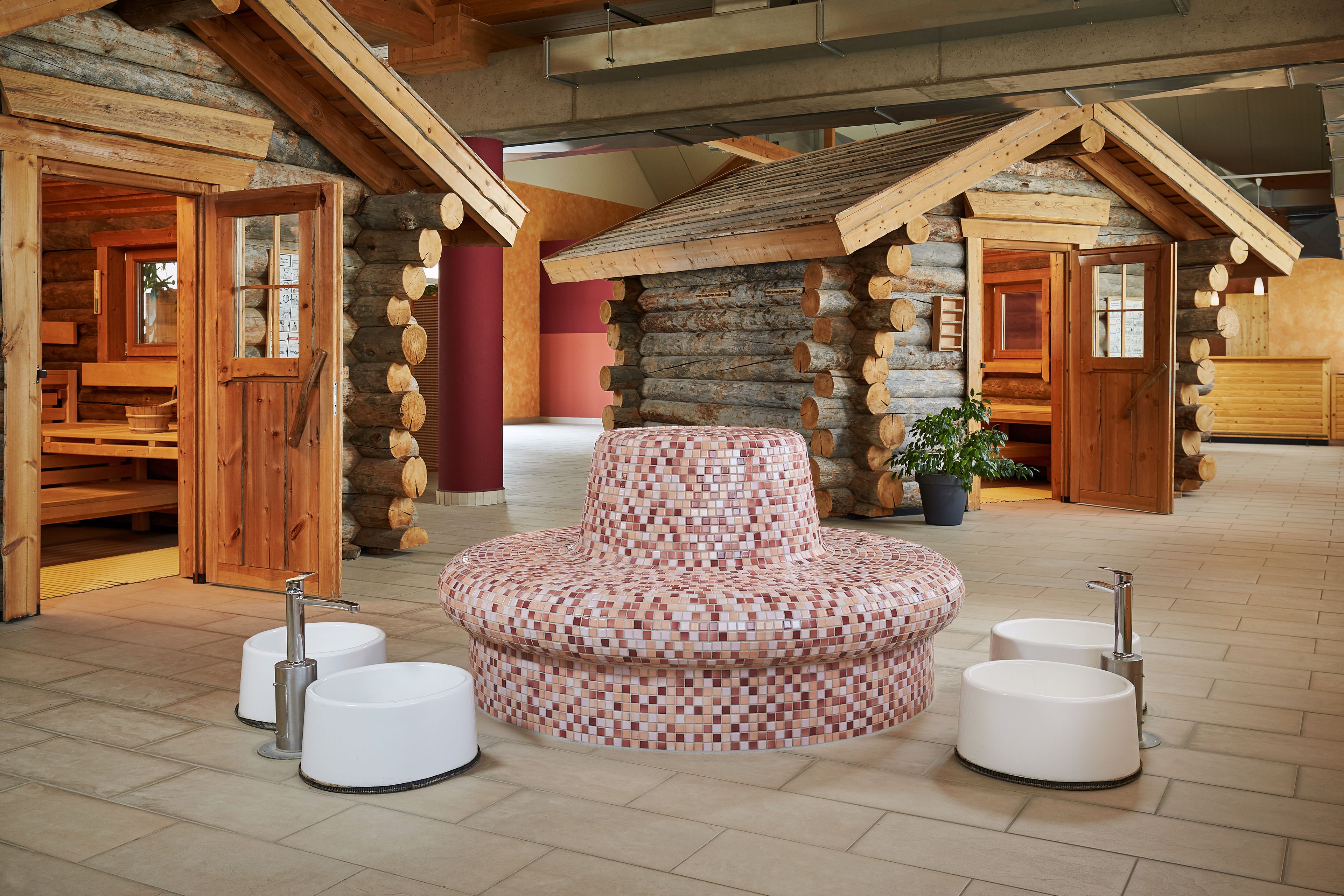 A log cabin interior with a central mosaic bench, two sinks, and wooden walls.