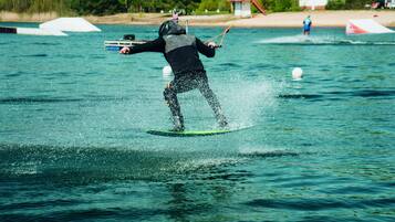 Uma pessoa praticando wakeboard em um corpo d'água, com céu claro e árvores ao fundo.