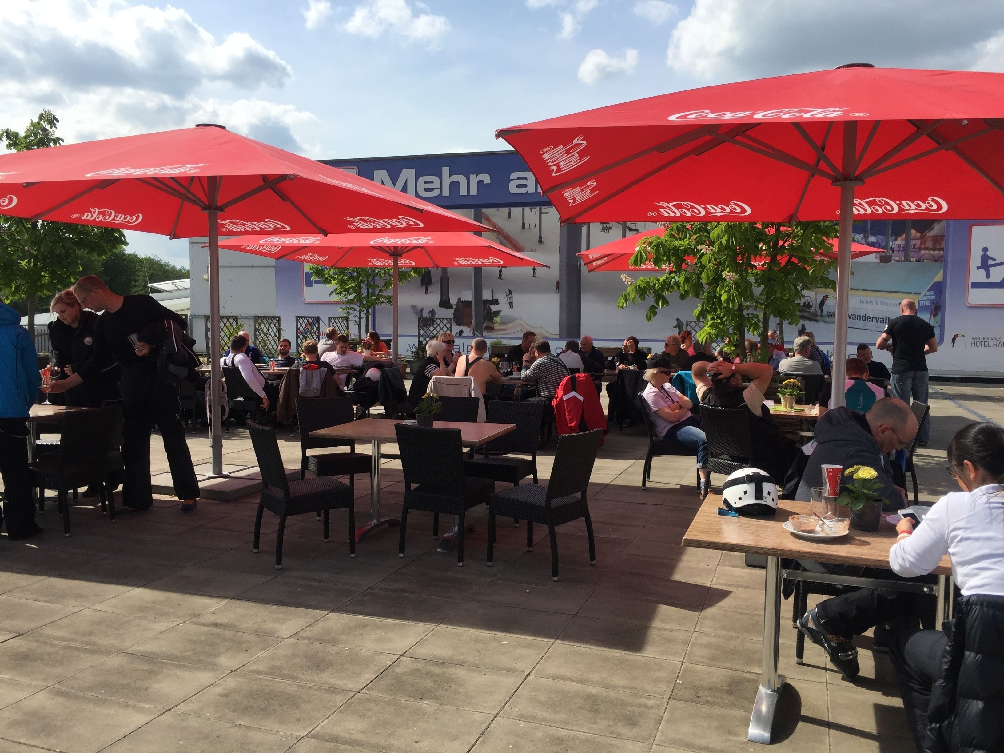 An outdoor seating area with tables and chairs under red umbrellas, a blue sign in the background, and people dining.