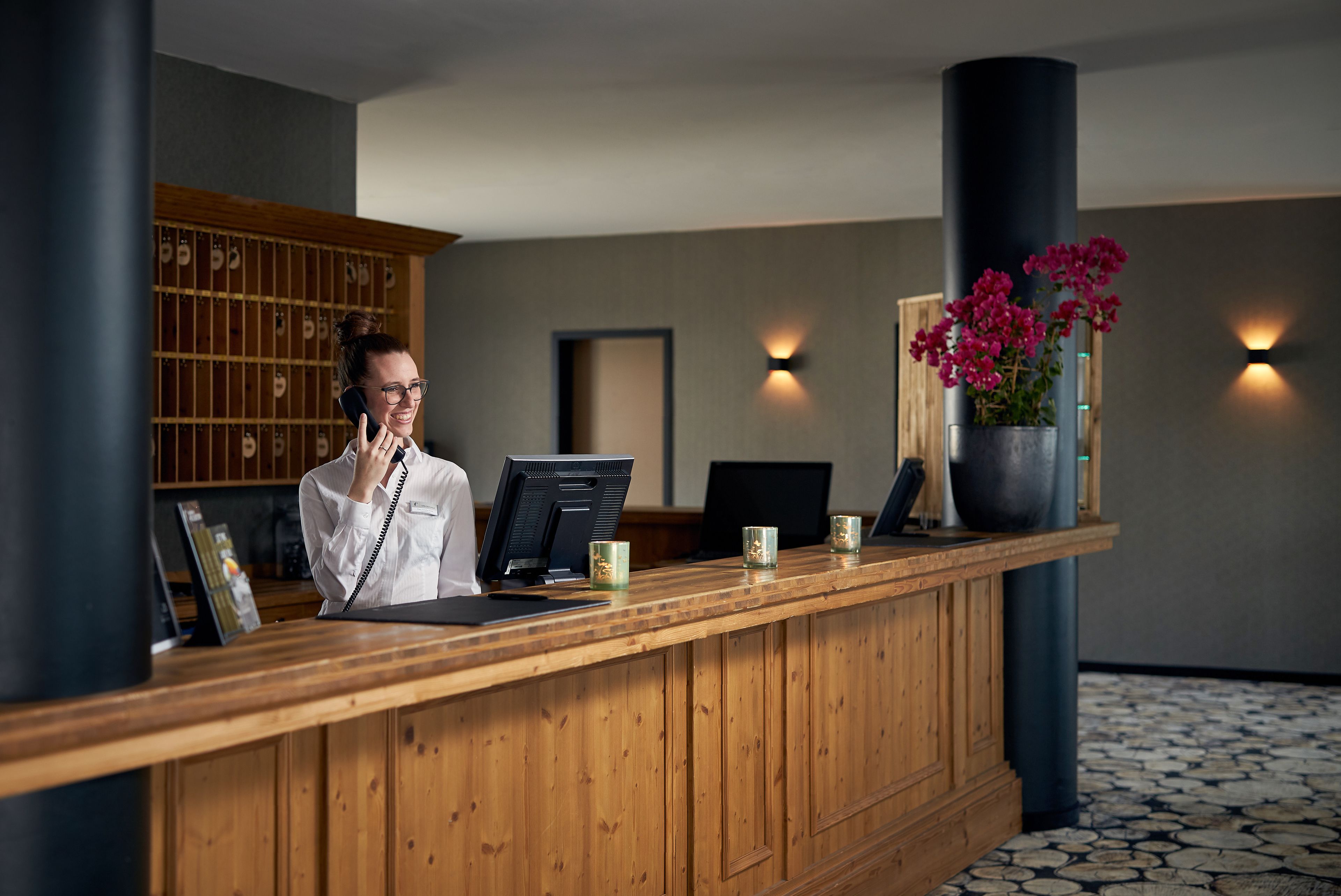 A wooden reception desk with a computer monitor, a telephone, and a vase with flowers.