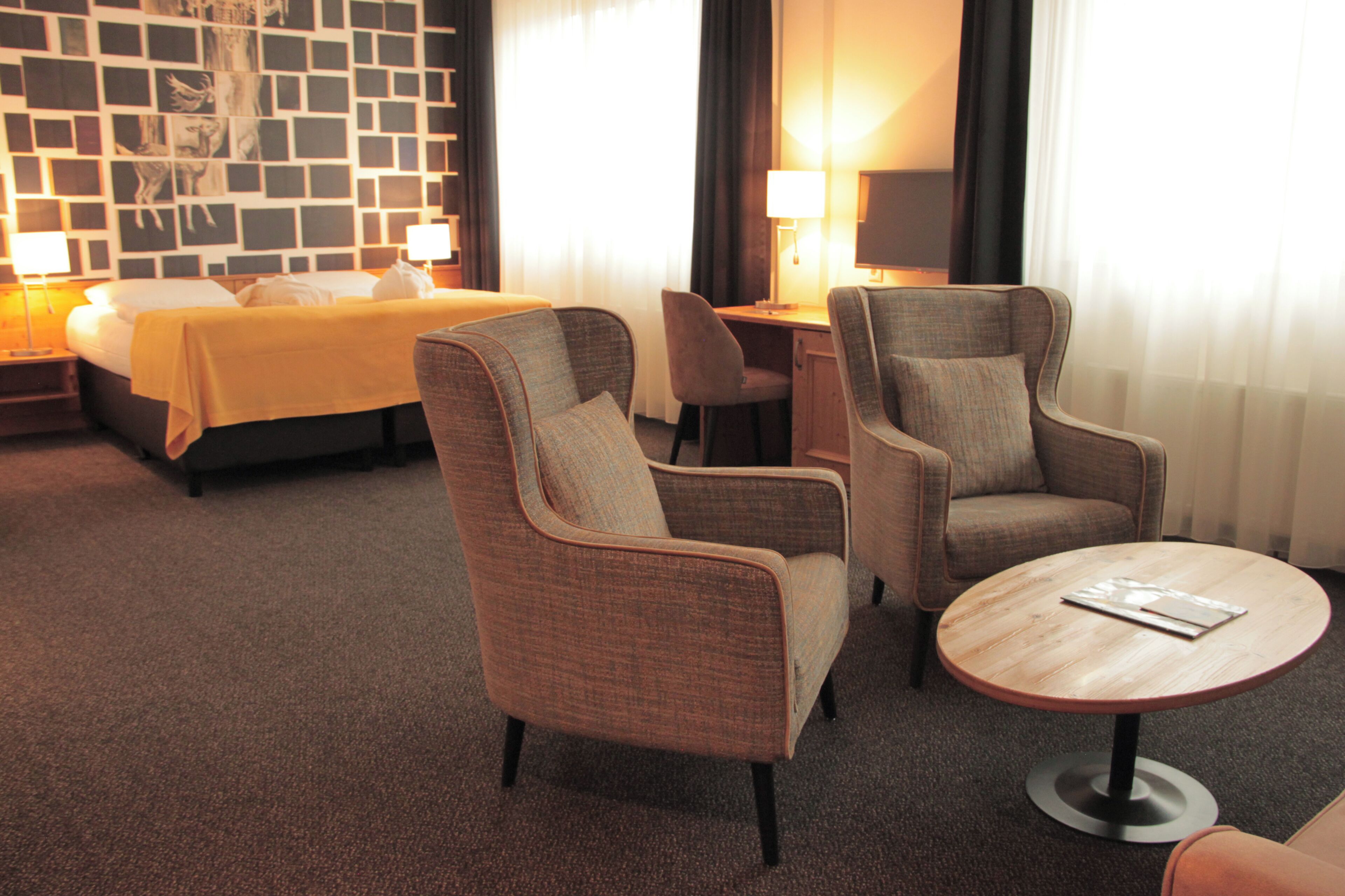 A hotel room with a bed, two armchairs, a small table, and a desk with a lamp.