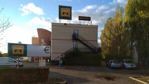 A hotel with a 'BIENVENUE' sign, a staircase leading to the entrance, and a parking area with cars. - hotelF1 Maurepas (Maurepas)