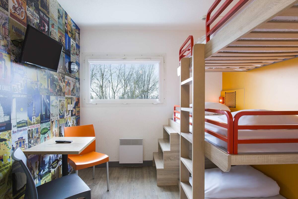 Une chambre compacte comprenant un lit superposé, un bureau avec une chaise, une fenêtre donnant sur la verdure et un mur décoré d’affiches de voyage.