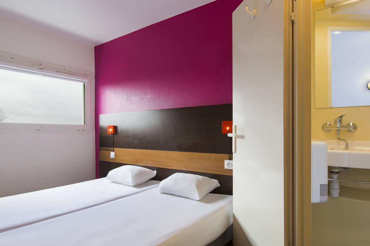 Une chambre d’hôtel comprenant un lit, une table de chevet, une lampe fixée au mur et une salle de bain avec un lavabo et un miroir.