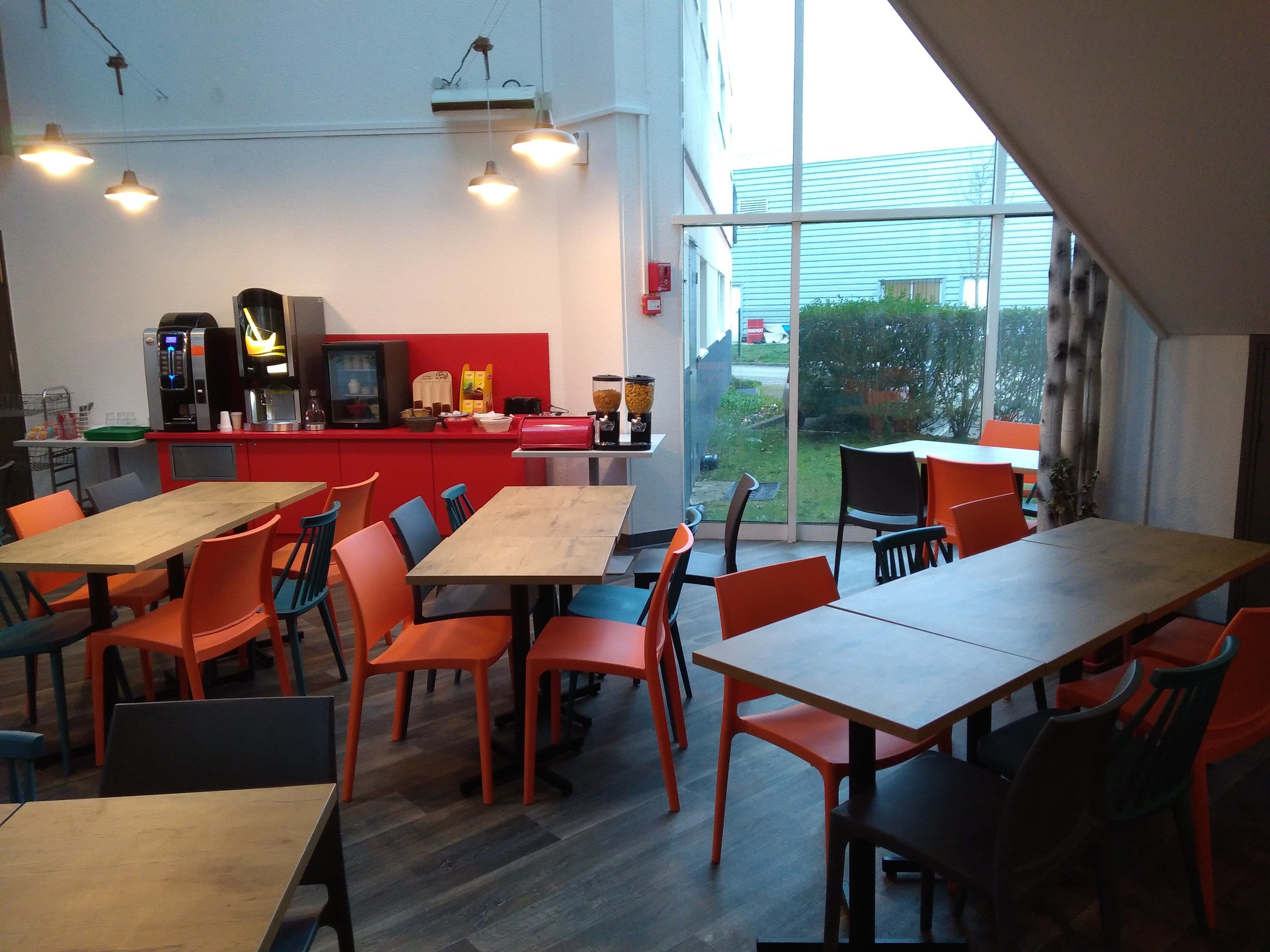 Un café moderne avec des tables en bois et des chaises orange, un comptoir rouge avec des machines à café, et de grandes fenêtres donnant sur l’extérieur.