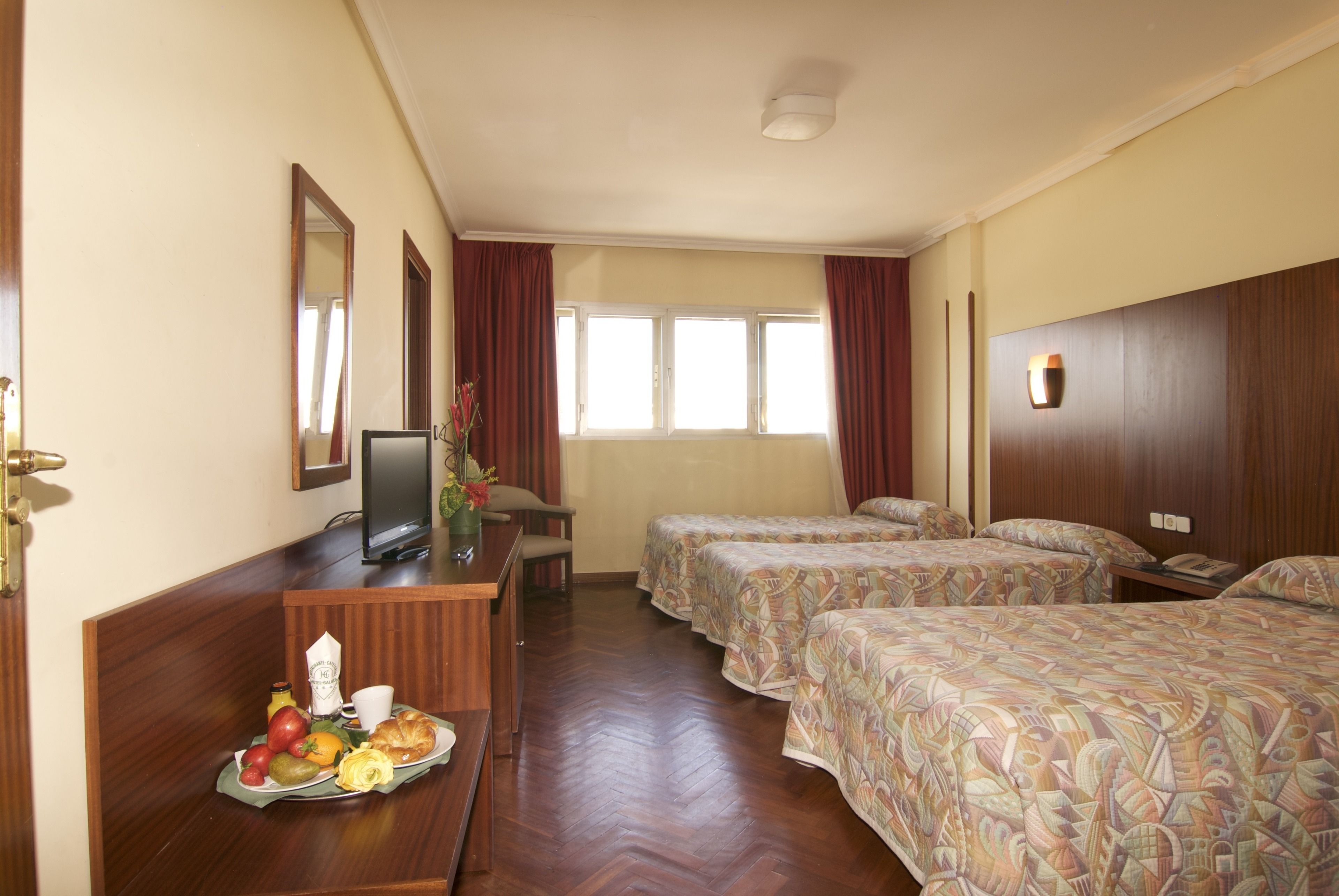 Een hotelkamer met een houten bureau, een televisie, twee bedden met beddengoed met een patroon, een raam met gordijnen en een houten hoofdeinde.