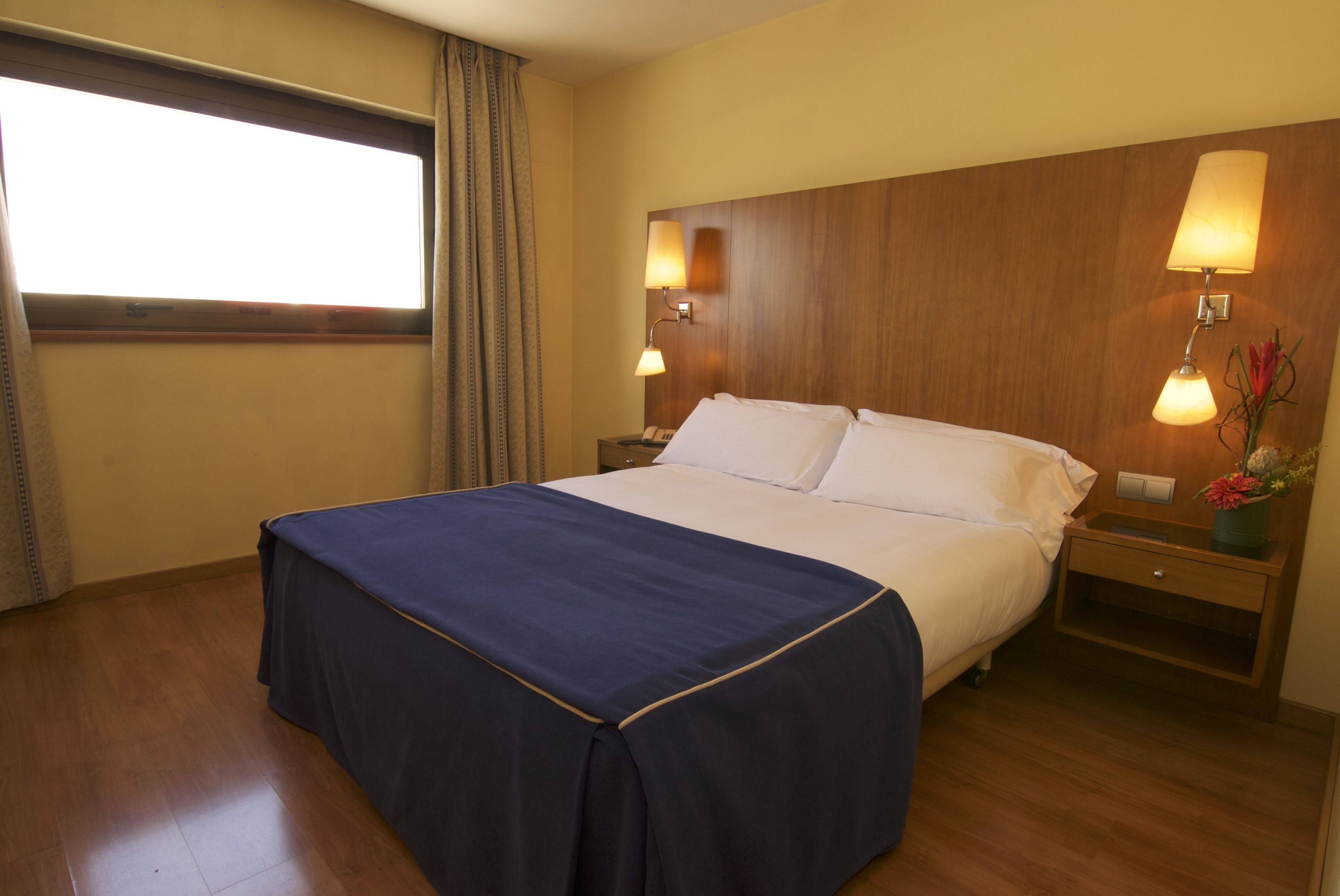 Habitación de hotel con una cama grande, dos mesitas de noche con lámparas, una ventana con cortinas y un cabecero de madera.
