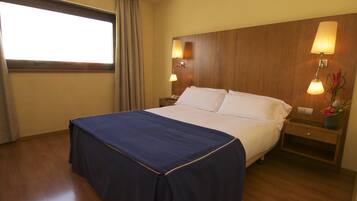 Habitación de hotel con una cama grande, dos mesitas de noche con lámparas, una ventana con cortinas y un cabecero de madera.